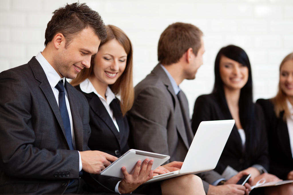 Man in black suit showing something on his tablet to collogue who is working on laptop