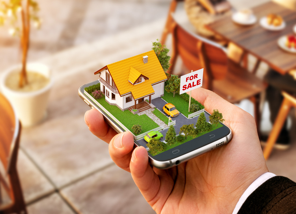A person holding a phone with a 3D model of a house with a For Sale sign