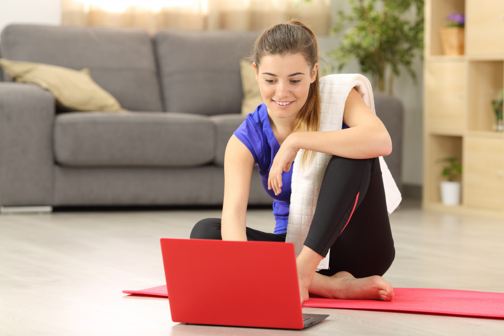 A woman using a laptop while sitting on a yoga mat on the floor at home