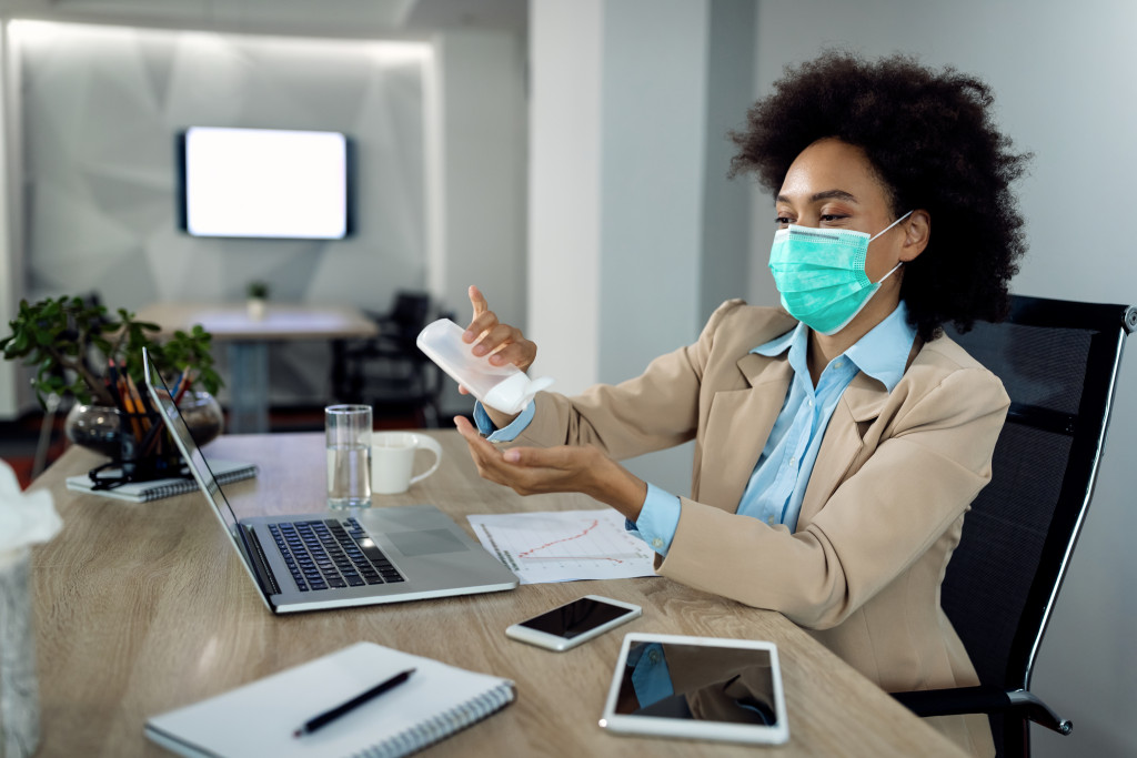 American businesswoman using hands sanitizer and wearing face mask while working in the office during coronavirus pandemic.