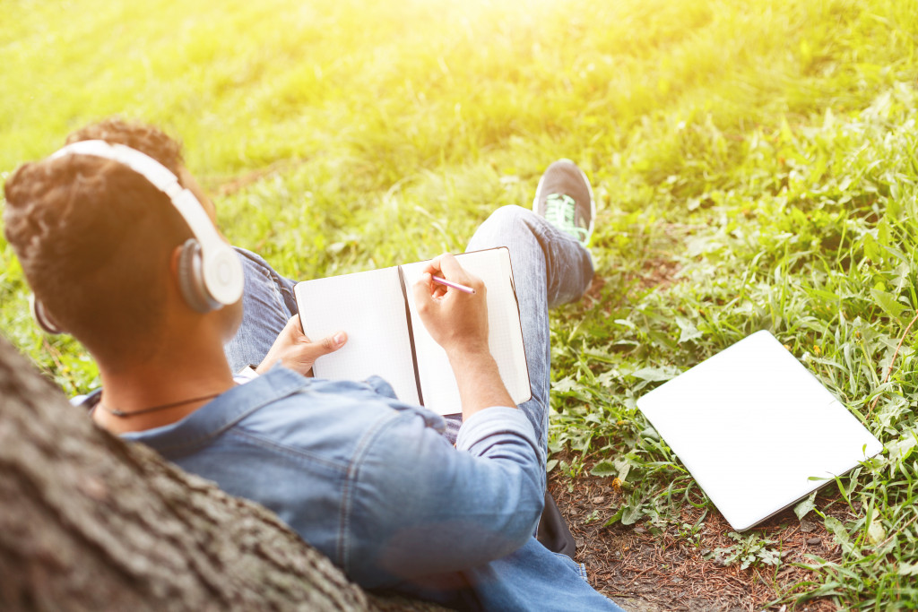 man writing in journal while wearing headphones seated next to a tree