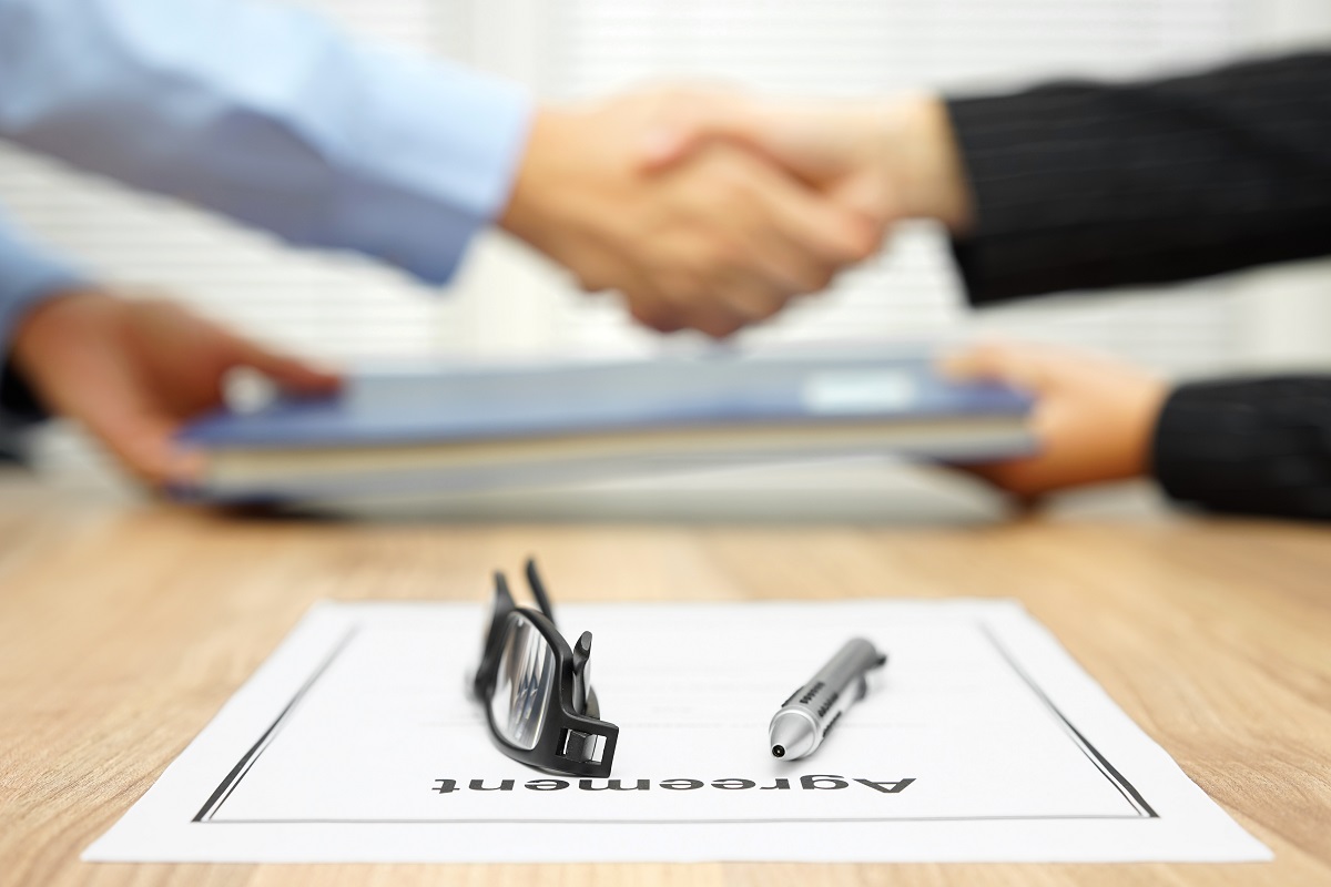 Two business people shaking hands in the background with pen and glasses on a paper agreement